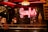 A warmly lit interior featuring a neon sign that reads 'Gaufres'. The setting includes a display of waffles and an ice cream cone. There is a menu written on a blackboard in the background alongside posters. The ambiance is cozy with a vintage feel, accented by a patterned wall and hanging lamps.