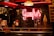 A warmly lit interior featuring a neon sign that reads 'Gaufres'. The setting includes a display of waffles and an ice cream cone. There is a menu written on a blackboard in the background alongside posters. The ambiance is cozy with a vintage feel, accented by a patterned wall and hanging lamps.