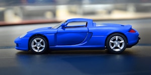 A blue toy car is situated on a flat surface, with a focus on its sleek design and smooth lines. The car features metallic silver wheels and an overall sporty appearance, resembling a convertible sports car model.