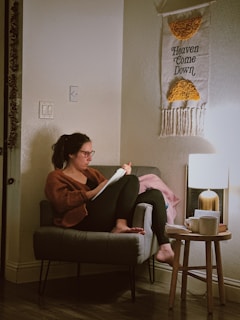 A person with glasses is sitting on a chair in a cozy room, reading or writing on paper. The individual is surrounded by a warm light and a tasseled wall hanging with the words 'Heaven Come Down.' A small table beside the chair holds a lamp, a stack of books, and a cup.