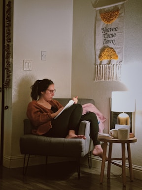 A person with glasses is sitting on a chair in a cozy room, reading or writing on paper. The individual is surrounded by a warm light and a tasseled wall hanging with the words 'Heaven Come Down.' A small table beside the chair holds a lamp, a stack of books, and a cup.