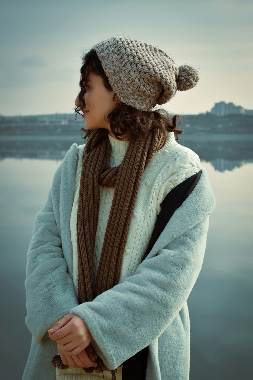 a woman standing next to a body of water wearing a hat and scarf