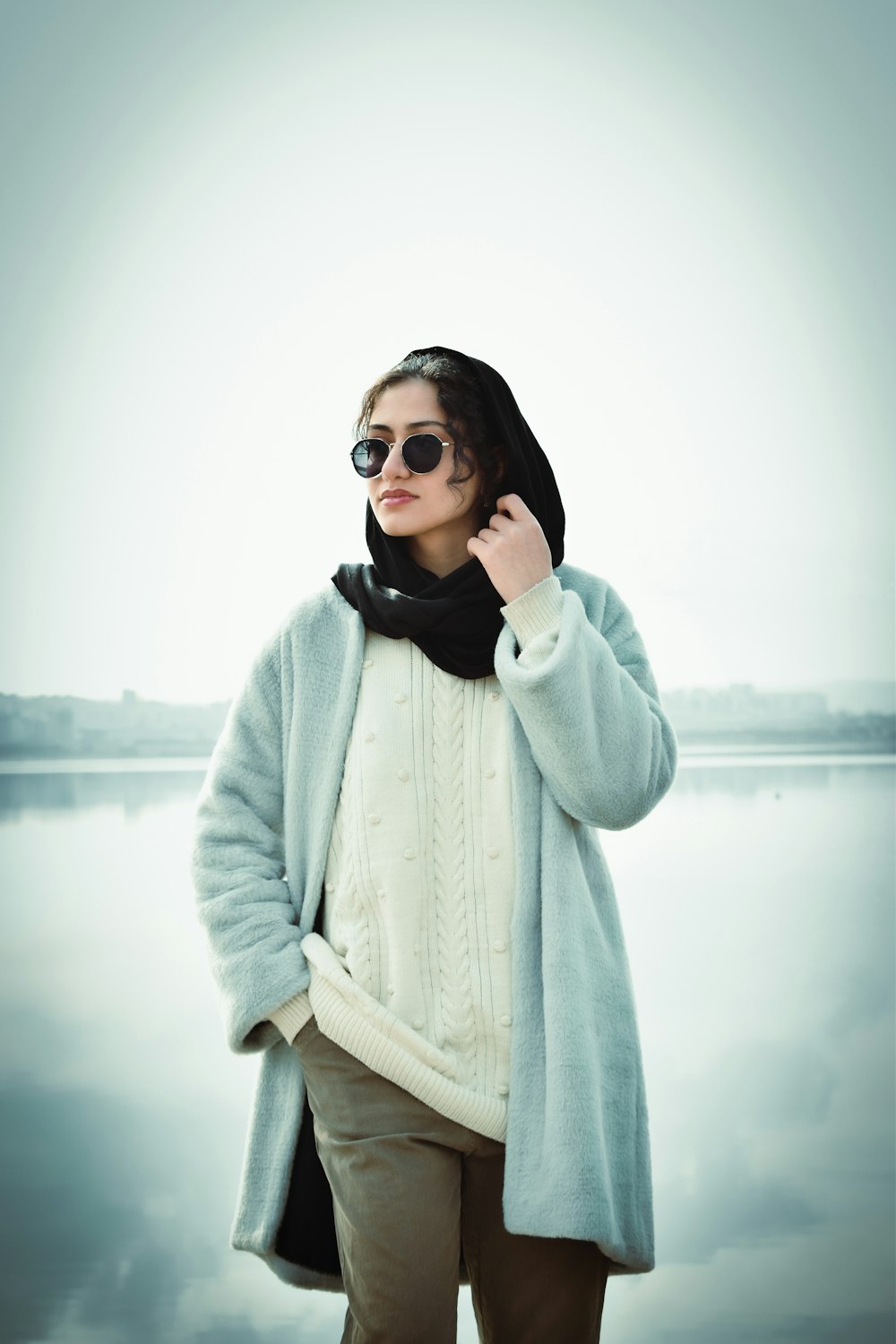 a woman wearing sunglasses and a coat by the water