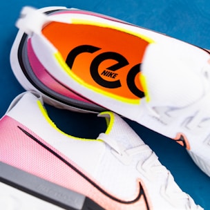 A pair of white athletic shoes with pink gradients and neon yellow accents. The inner sole is vibrant orange with bold, black text 'Nike' visible. The shoes are placed on a blue surface, showcasing a dynamic and modern design.