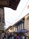 A bustling street scene with a mix of locals and tourists walking along a narrow walkway. The street is lined with shops displaying signs such as 'Sunshine India' and 'House of Cashmere'. Vibrant clothing and textiles are visible in shop displays. The buildings have an old architectural style with sloping roofs and are painted in earthy tones. Overhead wires crisscross the sky, adding to the atmosphere of a lively market area.