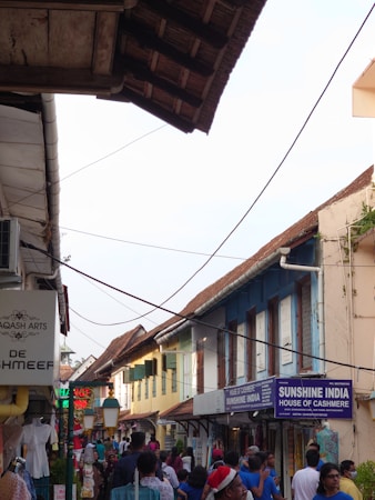 A bustling street scene with a mix of locals and tourists walking along a narrow walkway. The street is lined with shops displaying signs such as 'Sunshine India' and 'House of Cashmere'. Vibrant clothing and textiles are visible in shop displays. The buildings have an old architectural style with sloping roofs and are painted in earthy tones. Overhead wires crisscross the sky, adding to the atmosphere of a lively market area.