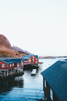 A picturesque scene featuring several red wooden cabins built on stilts along a calm body of water, with a mountainous background. The cabins have white window frames and dark roofs, creating a striking contrast with the surrounding landscape. Reflections are visible in the water, and the sky is clear.