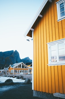 A vibrant yellow wooden building with white trim is set against a backdrop of rugged mountains and a clear sky. The architecture includes large windows and a gabled roof, suggesting a cozy, rustic setting. Nearby, other similar structures are visible, surrounded by rocks and a chain fence, with a hint of frost on the ground.
