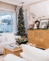 A cozy living room decorated for the holidays, featuring a small Christmas tree adorned with ornaments beside a window. The room includes a large wooden cabinet with decorative items like a lamp, framed artwork with 'joy' written on it, and greenery. A soft white couch with cushions and a wooden coffee table topped with books and a festive floral arrangement completes the setting.