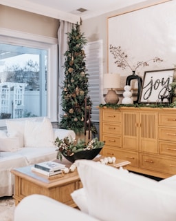 A cozy living room decorated for the holidays, featuring a small Christmas tree adorned with ornaments beside a window. The room includes a large wooden cabinet with decorative items like a lamp, framed artwork with 'joy' written on it, and greenery. A soft white couch with cushions and a wooden coffee table topped with books and a festive floral arrangement completes the setting.