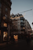 A Parisian street scene at dusk featuring classic Haussmann-style buildings with ornate architecture. Warm streetlights and glowing shop windows illuminate the street, and a neon sign displays the word 'Montorgueil' in cursive. People walk along the dimly lit street, and the sky is a soft shade of blue, indicating early evening.