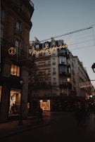 A Parisian street scene at dusk featuring classic Haussmann-style buildings with ornate architecture. Warm streetlights and glowing shop windows illuminate the street, and a neon sign displays the word 'Montorgueil' in cursive. People walk along the dimly lit street, and the sky is a soft shade of blue, indicating early evening.