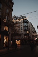A Parisian street scene at dusk featuring classic Haussmann-style buildings with ornate architecture. Warm streetlights and glowing shop windows illuminate the street, and a neon sign displays the word 'Montorgueil' in cursive. People walk along the dimly lit street, and the sky is a soft shade of blue, indicating early evening.