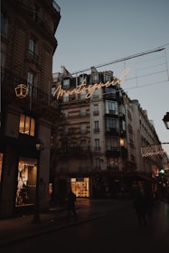 A Parisian street scene at dusk featuring classic Haussmann-style buildings with ornate architecture. Warm streetlights and glowing shop windows illuminate the street, and a neon sign displays the word 'Montorgueil' in cursive. People walk along the dimly lit street, and the sky is a soft shade of blue, indicating early evening.
