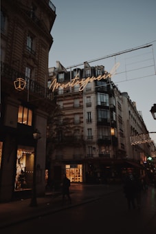 A Parisian street scene at dusk featuring classic Haussmann-style buildings with ornate architecture. Warm streetlights and glowing shop windows illuminate the street, and a neon sign displays the word 'Montorgueil' in cursive. People walk along the dimly lit street, and the sky is a soft shade of blue, indicating early evening.