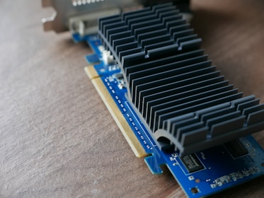 A computer graphics card rests on a wooden surface, showcasing intricate metallic heat sinks and a blue circuit board. The card features various electronic components and gold connectors for interfacing with a computer.
