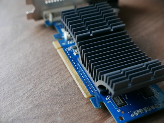A computer graphics card rests on a wooden surface, showcasing intricate metallic heat sinks and a blue circuit board. The card features various electronic components and gold connectors for interfacing with a computer.