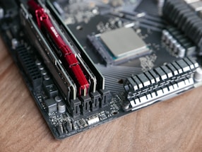 A close-up of a computer motherboard with visible RAM sticks and a CPU socket on a wooden surface. The RAM sticks are red, and various components and circuitry are visible on the board, including heat sinks and capacitors.