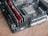 A close-up of a computer motherboard with visible RAM sticks and a CPU socket on a wooden surface. The RAM sticks are red, and various components and circuitry are visible on the board, including heat sinks and capacitors.