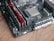A close-up of a computer motherboard with visible RAM sticks and a CPU socket on a wooden surface. The RAM sticks are red, and various components and circuitry are visible on the board, including heat sinks and capacitors.