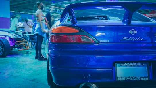 A vibrant scene in a parking garage features the rear end of a blue Nissan Silvia car in the foreground. The area is bustling with activity, as people move around a display of modified cars with colorful lighting, adding a dynamic, lively atmosphere.