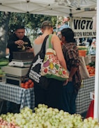 A farmer's market scene with people standing near a stall. A person in a black hoodie is handling a scale behind a table covered with a blue checkered cloth, and there are various fruits like tomatoes and grapes displayed in the foreground and background. Another person with a tote bag and tank top stands with their back to the camera.