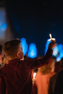 A young person wearing a red plaid shirt holds up a candle in a dark setting, surrounded by other individuals in soft focus. The background features blue bokeh lights, creating a serene and peaceful atmosphere.