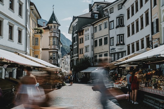 The scene captures a lively and bustling street market located between a row of European-style buildings. People are blurred as they walk by, emphasizing motion and activity. The buildings feature unique and intricate architecture, with balconies and detailed facades. The market stalls are covered with awnings and display a variety of goods, attracting locals and tourists alike.