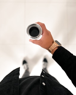 A person holds a stainless steel travel mug filled with a dark liquid, possibly coffee. The person is wearing a dark long-sleeve shirt with white buttons, a gold or rose gold metal watch, and black and white shoes visible at the bottom of the image. The background is a plain, light-colored surface.