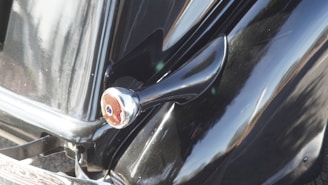 Close-up of a classic car's glossy black body, featuring a prominent silver side mirror and smooth curves, reflecting sunlight.