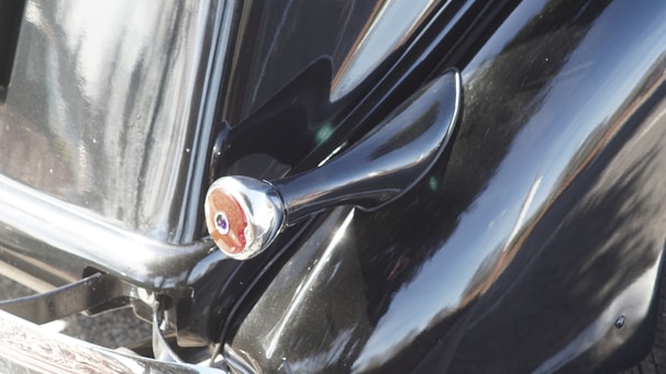 Close-up of a classic car's glossy black body, featuring a prominent silver side mirror and smooth curves, reflecting sunlight.