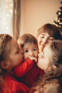 Three children are close together, with two older girls kissing the cheeks of a younger baby who is held in the center. The setting appears warm and cozy with soft lighting and a blurred background featuring a Christmas tree with lights.
