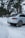 A silver luxury SUV is parked on a snow-covered ground, surrounded by tall pine trees. The vehicle's rear view is visible, showing sporty wheels and a sleek design.