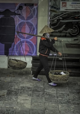 A person wearing a traditional conical hat carries baskets suspended from a pole over their shoulder. The baskets are made of a woven material and appear to contain items covered by a cloth. The person is walking on a tiled sidewalk in front of a storefront displaying motorbikes and advertisements.