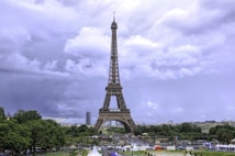 The image features the Eiffel Tower, prominently standing tall against a backdrop of cloudy skies. There is lush greenery surrounding the monument and various structures and pathways can be seen at the base of the tower. People and vehicles are visible, adding a dynamic element to the scenic view.