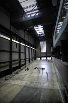 A large, industrial-style interior space with an expansive, empty concrete floor. The high ceiling features exposed steel beams and large skylights, allowing natural light to filter in. On the left side, a series of balconies and railings can be seen, while the far end of the room has a tall window with vertical bars. A group of people, including individuals and small clusters, are dispersed across the floor, walking or sitting.