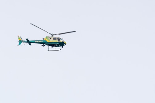 A green and yellow helicopter marked with the word 'Sheriff' is flying in a clear blue sky. The helicopter's rotors are spinning and it has a tail rotor for stabilization.