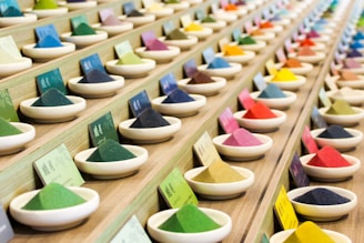 Multiple rows of small ceramic bowls filled with vibrantly colored powders are neatly arranged on a wooden display. Each bowl is accompanied by a card, possibly indicating the name or type of the powder. The arrangement is orderly and aesthetically pleasing.