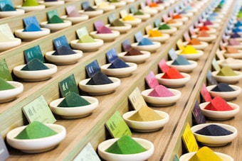 Multiple rows of small ceramic bowls filled with vibrantly colored powders are neatly arranged on a wooden display. Each bowl is accompanied by a card, possibly indicating the name or type of the powder. The arrangement is orderly and aesthetically pleasing.