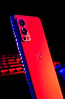 A sleek smartphone with a matte finish is highlighted by intense red and blue lighting. The device features a noticeable rear camera module with three lenses and a flash. In the background, there are blurred red and blue illuminated components possibly indicating a tech or gaming setup.