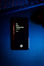 A smartphone displaying the time as five twenty-four, with a fingerprint icon at the bottom, lies on a dark surface. Above the phone, part of a keyboard is visible, illuminated by blue and red lighting.
