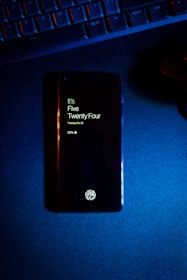 A smartphone displaying the time as five twenty-four, with a fingerprint icon at the bottom, lies on a dark surface. Above the phone, part of a keyboard is visible, illuminated by blue and red lighting.