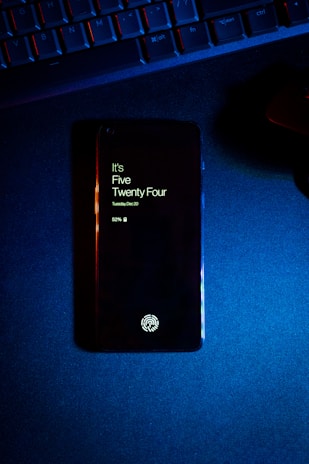 A smartphone displaying the time as five twenty-four, with a fingerprint icon at the bottom, lies on a dark surface. Above the phone, part of a keyboard is visible, illuminated by blue and red lighting.