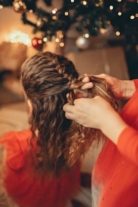A person is braiding a child's hair, with a focus on the intricate layers of the braid. The background features blurred holiday decorations, including a Christmas tree with lights and ornaments.