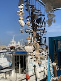 A display of various handmade nautical decorations and ornaments, including wooden fish and seashells, hung together with a small sign labeled 'Rhodes.' In the background, a marina with several boats and ships is visible under a clear blue sky.