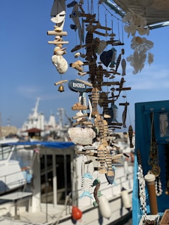 A display of various handmade nautical decorations and ornaments, including wooden fish and seashells, hung together with a small sign labeled 'Rhodes.' In the background, a marina with several boats and ships is visible under a clear blue sky.