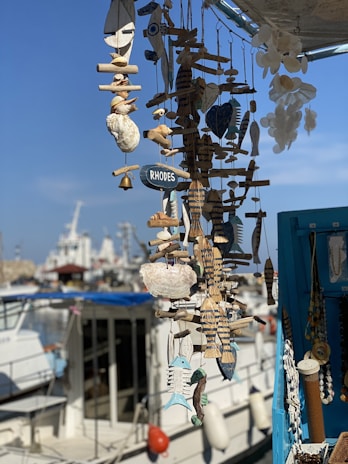 A display of various handmade nautical decorations and ornaments, including wooden fish and seashells, hung together with a small sign labeled 'Rhodes.' In the background, a marina with several boats and ships is visible under a clear blue sky.