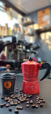 A red speckled stovetop espresso maker is placed on a black surface alongside a black coffee cup with a logo. Coffee beans are scattered around the espresso maker and cup. The background features a blurred coffee shop setting with various equipment and a warm-toned ambiance.