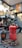 A red speckled stovetop espresso maker is placed on a black surface alongside a black coffee cup with a logo. Coffee beans are scattered around the espresso maker and cup. The background features a blurred coffee shop setting with various equipment and a warm-toned ambiance.