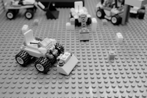 A monochrome image displaying a small model vehicle made of interlocking plastic bricks on a textured baseplate. Several other brick constructs and scattered bricks are visible in the background.
